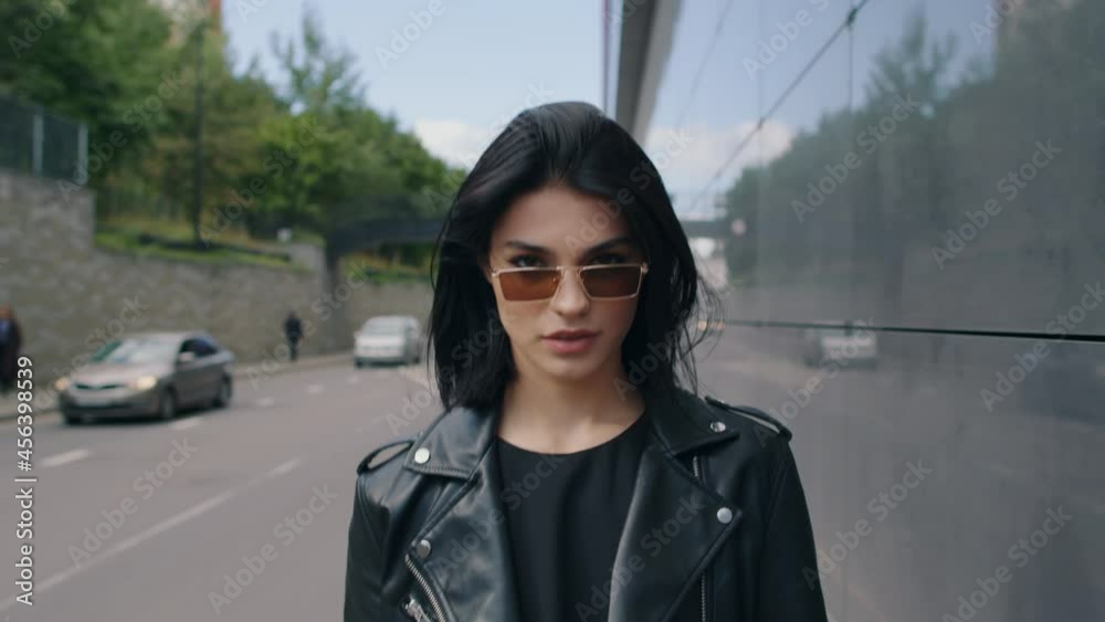 Confident seductive happy brunette female in stylish look black leather jacket and sunglasses walking outdoors along city street looking at camera flirts smiling on sunny day. Urban style fashion Confident seductive happy brunette female in stylish look black leather jacket and sunglasses walking outdoors along city street looking at camera flirts smiling on sunny day. Urban style fashion
