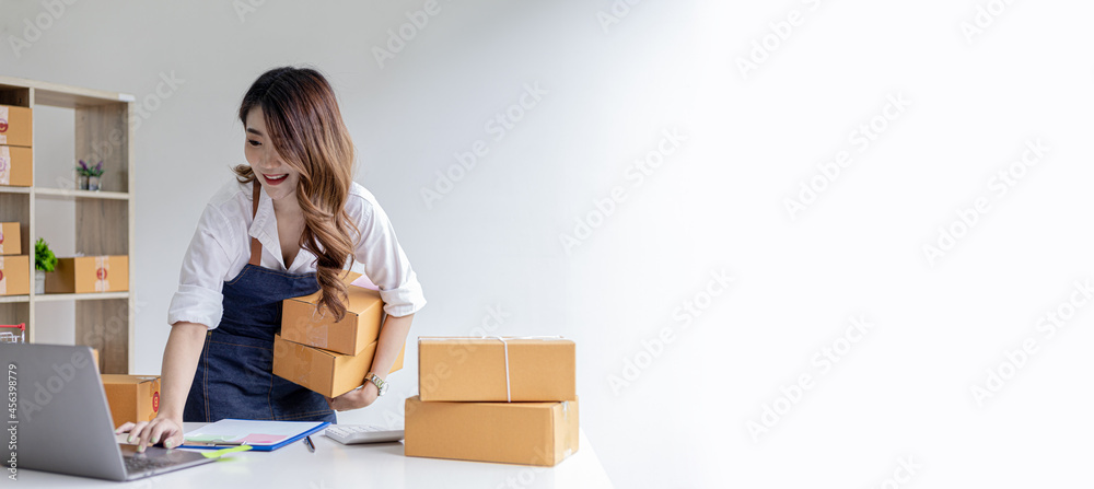 Asian woman holding parcel boxes and checking orders from laptop, she owns an online store, she packs and ships through a private transport company. Online selling and online shopping concepts. Asian woman holding parcel boxes and checking orders from laptop, she owns an online store, she packs and ships through a private transport company. Online selling and online shopping concepts.