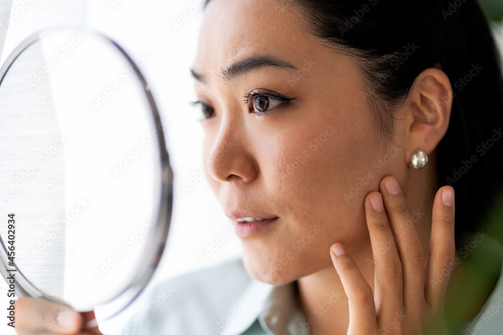 Young Asian woman looking herself in the mirror. Worried about acne caused by wearing a mask. Maskne. Problems with acne. Young Asian woman looking herself in the mirror. Worried about acne caused by wearing a mask. Maskne. Problems with acne.