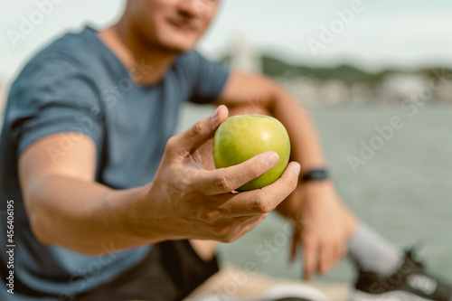 Close up sport man is sitting on cliff near sea and eating green apple after morning cardio.