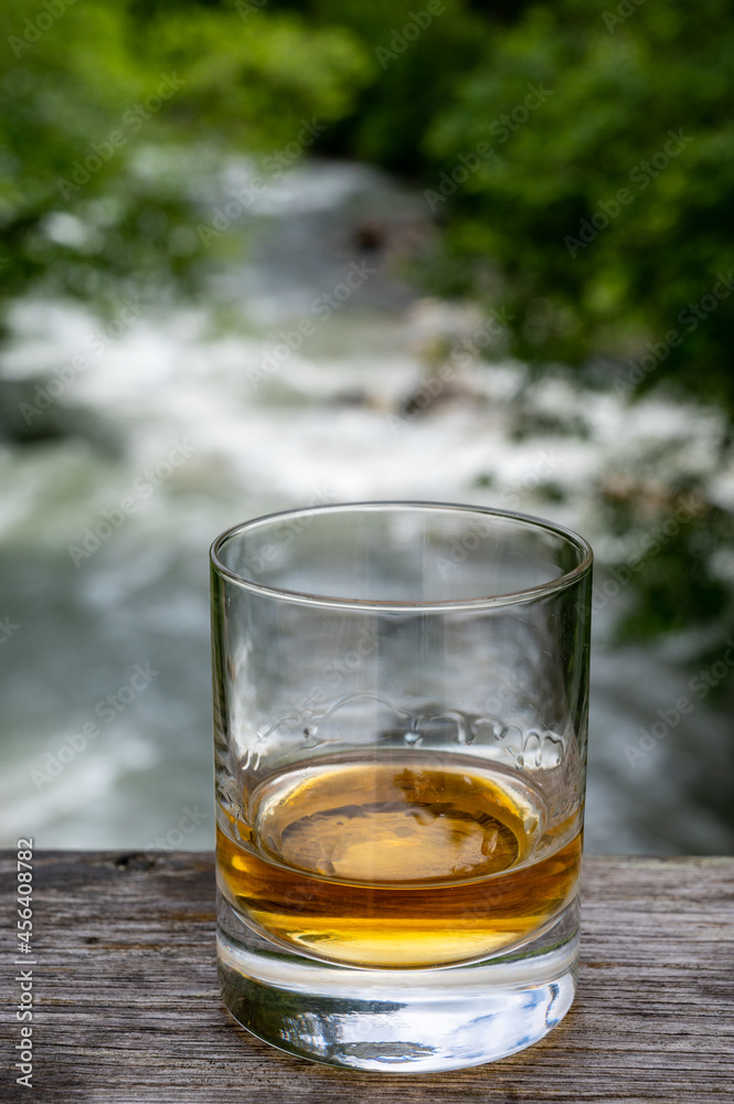 Glass of strong scotch single malt whisky with fast flowing mountain river on background Glass of strong scotch single malt whisky with fast flowing mountain river on background
