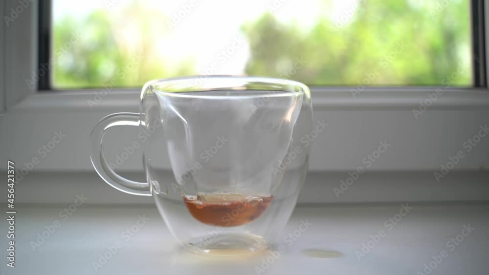Person pours tea from teapot into glass cup standing on white windowsill and spills few drops past, takes napkin and wipes table from splashes. Morning ceremony. Person pours tea from teapot into glass cup standing on white windowsill and spills few drops past, takes napkin and wipes table from splashes. Morning ceremony.