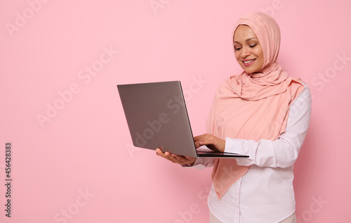 Portrait of a beautiful Muslim Arab woman in pink hijab working on laptop, isolated on colored background with copy space. Successful programmer, IT female worker, start-up, business lady