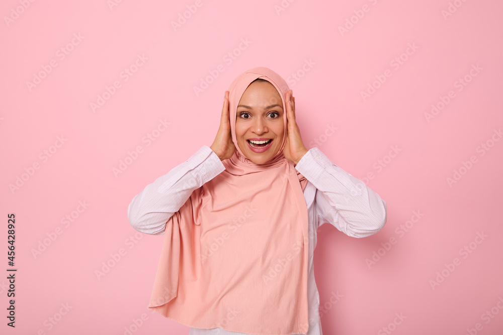 Happy religious Muslim woman in pink hijab holding her head with hands and smiling with toothy smile looking at camera with surprised look, isolated on colored background with copy space. Happy religious Muslim woman in pink hijab holding her head with hands and smiling with toothy smile looking at camera with surprised look, isolated on colored background with copy space.