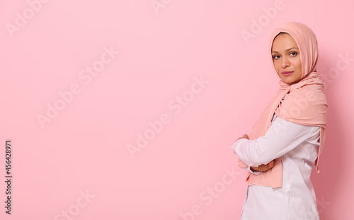 Confident portrait of Arabic muslim beautiful woman with attractive look and gaze, wearing a pink hijab and standing sideways against colored background with copy space