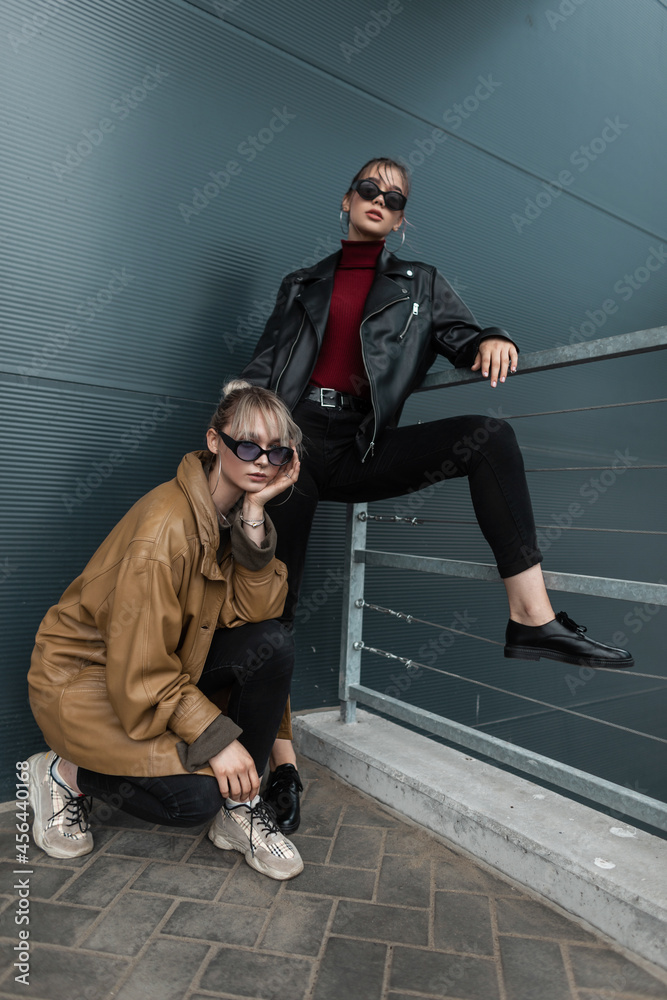 Two beautiful young women in trendy denim clothes with a leather jacket in sunglasses posing near a metal wall outdoors