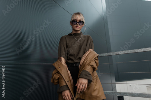 Fashionable model girl with cool sunglasses in a brown black jacket with black jeans and a sweater stands near a metal wall on the street
