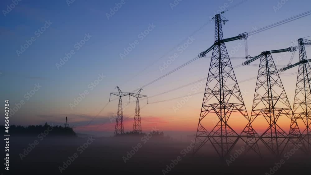 Aerial view high voltage steel power pylons in field covered with fog countryside. Misty early morning, dawn. Drone flight low over power transmission lines. Electric tower line, sunrise sunset