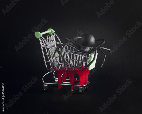 Minimal, dark, halloween bloody arrangement with big black spider in a supermarket cart with clotted blood dripping, isolated on the black background. Halloween decoration purchase concept. 