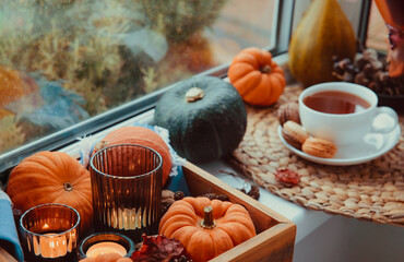 Autumn cozy composition. Pumpkins, candles, fall leaves on wooden tray and cup of hot tea with sweets on the windowsill. Autumn, fall, hygge home mood. Selective focus. Copy space