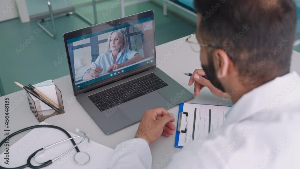 Videocall chat with female older patient and indian male doctor in modern clinic hospital using pc laptop computer, consulting online remotely. Telemedicine healthcare concept. Over shoulder view. Videocall chat with female older patient and indian male doctor in modern clinic hospital using pc laptop computer, consulting online remotely. Telemedicine healthcare concept. Over shoulder view.