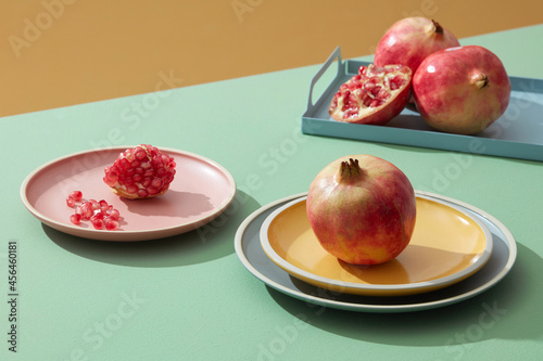 Close-up of pomegranate, half and seeds in pastel colored ceramic dishes on green table with shadow on background. Minimali