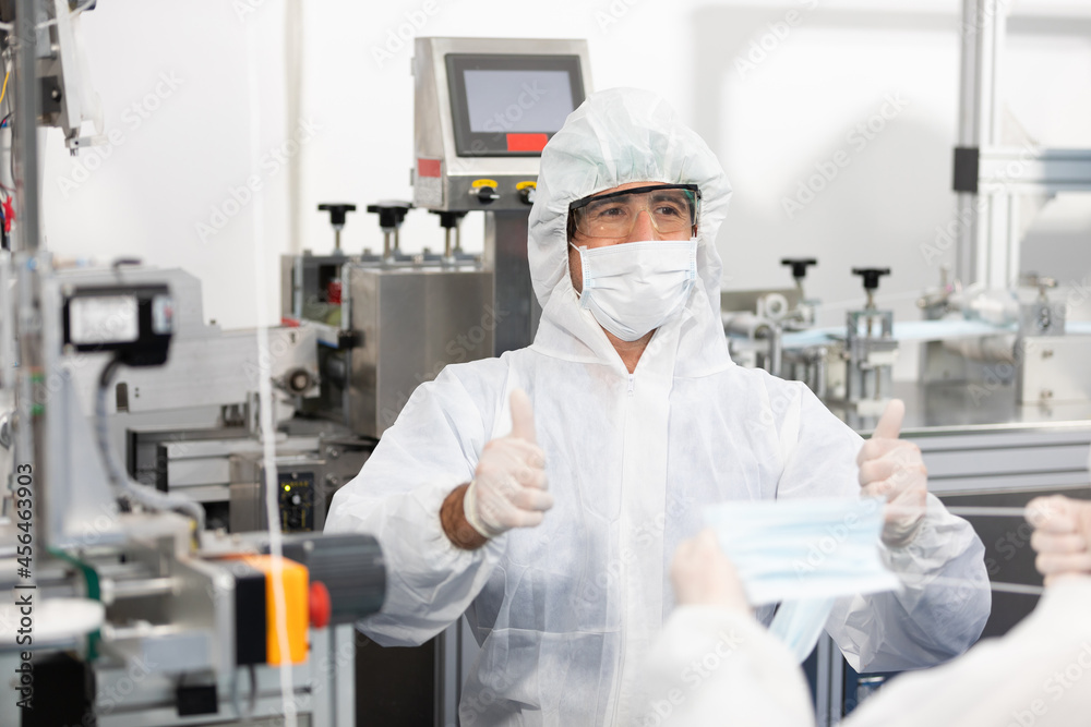 male engineers wearing personal protective equipment uniform(PPE) wearing medical face mask, producing medical face masks thumbs up pose beside machine in laboratory