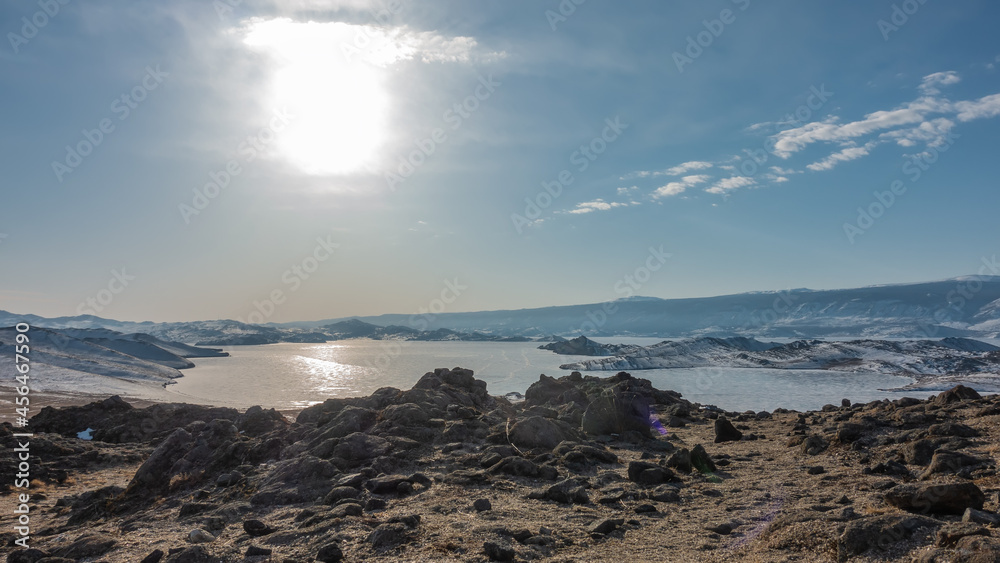Obraz premium The frozen lake is surrounded by snow-covered hills. The sun is shining in the blue sky. Glare on the ice. In the foreground is dry earth and rocks. Baikal