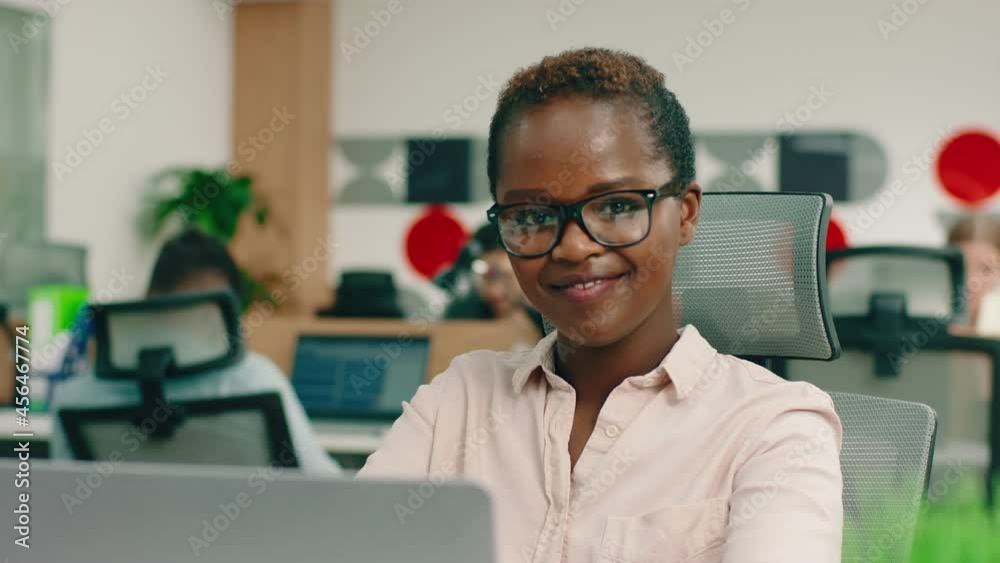 A very clever and charming young black woman with glasses is at her large office, sitting at the table and smiling greatly A very clever and charming young black woman with glasses is at her large office, sitting at the table and smiling greatly