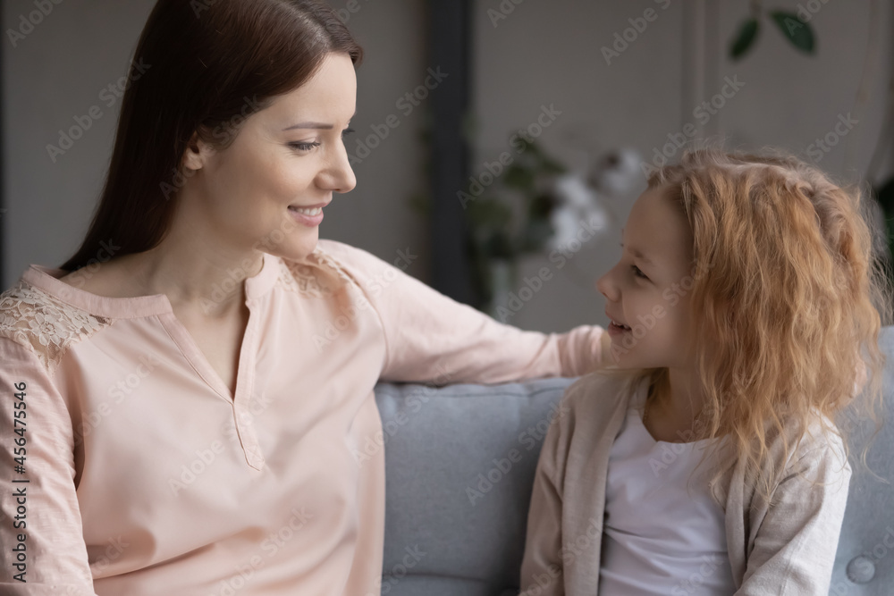 Smiling bonding young mother or nanny enjoying sincere conversation with cute small kid daughter, talking speaking communicating sitting on comfortable sofa in living room, trustful relations concept. Smiling bonding young mother or nanny enjoying sincere conversation with cute small kid daughter, talking speaking communicating sitting on comfortable sofa in living room, trustful relations concept.