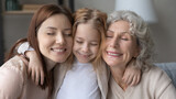Happy cute child girl cuddling affectionate young mother and elderly mature grandmother, enjoying sweet tender weekend time together. Smiling three generations family showing loving relations.