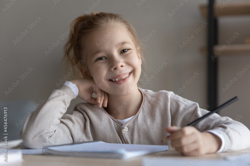 Head shot portrait of happy small preteen child girl sitting at table with pen in hands, enjoying doing school assignments, learning preparing for exams, handwriting notes, education concept. Head shot portrait of happy small preteen child girl sitting at table with pen in hands, enjoying doing school assignments, learning preparing for exams, handwriting notes, education concept.