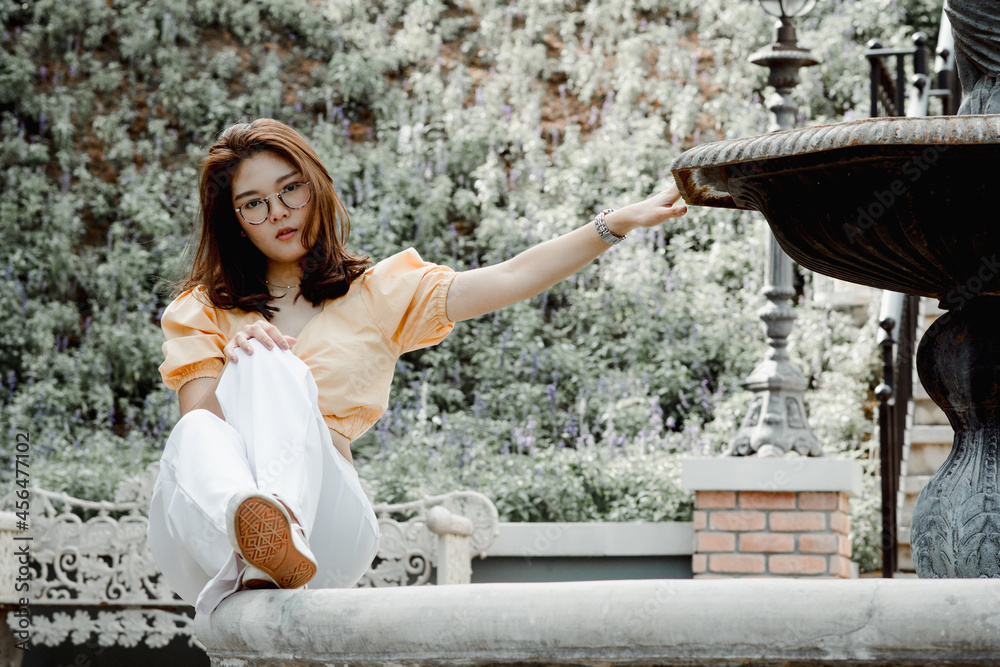 Beautiful young woman wearing glasses & yellow shirt and white pants. Travel on vacation. Posing for a photography, Sitting on the fountain, looking at the camera. The view behind is trees and nature.