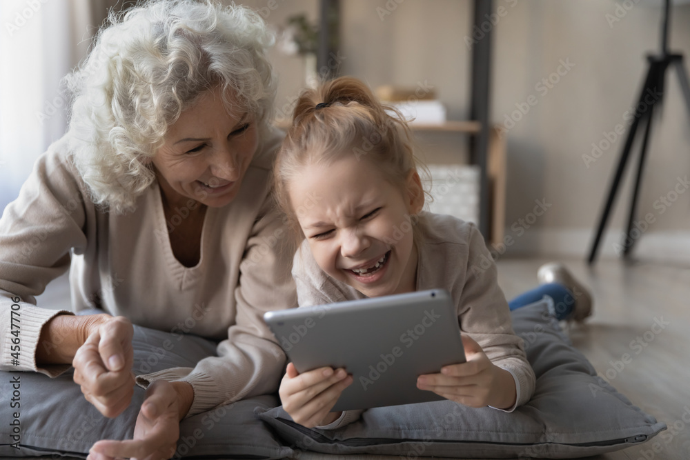 Addicted to modern technology happy small kid girl holding digital computer tablet, entertaining online laughing at funny photo video content with cheerful older granny, lying on floor on soft pillows