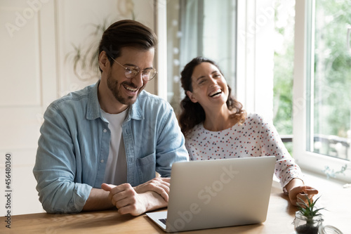 Fotografie Cheerful couple laughing sit at desk in front of laptop, enjoy funny humorous in