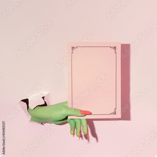 Creative layout with green painted hand holding pink paper copy space against pastel pink background. Halloween surreal idea. Minimal flat lay concept.