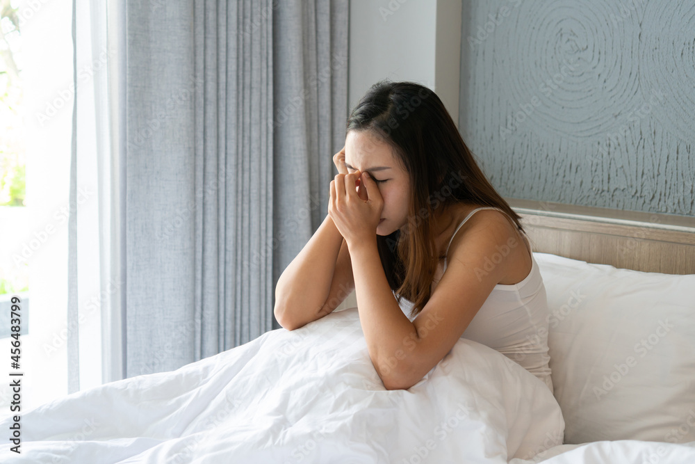 Stressed young Asian woman suffering on depression and sitting alone in bedroom at home. Girl have a headache from migraine sitting on bed. Sick, unhappy, disappointed concept Stressed young Asian woman suffering on depression and sitting alone in bedroom at home. Girl have a headache from migraine sitting on bed. Sick, unhappy, disappointed concept