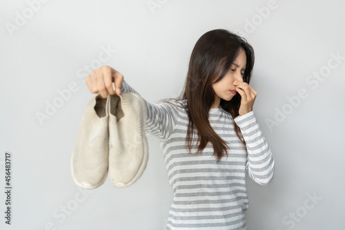 Young Asian woman is feeling disgusted with smell from shoes after long wearing. female holding dirty stinky shoes. Footwear needs in cleaning and odor removal. Unpleasant smell concept photo