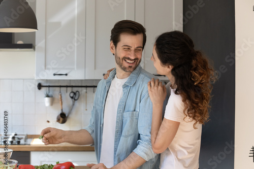 Happy homeowner couple in love cook together at home. Young wife hugs laughing husband standing in modern kitchen enjoy cookery on weekend. Healthy eating, romantic date, food delivery service concept