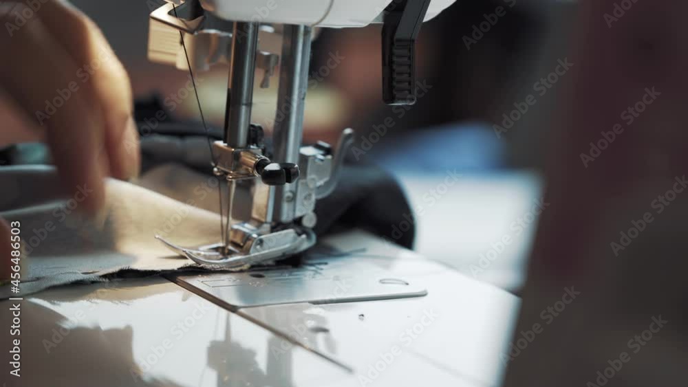 Close-up of foot of sewing machine making the seam with black thread. Women's fingers move fabric for stitching and pull needles out of material. Making clothes at home. Dressmaker's workshop. Close-up of foot of sewing machine making the seam with black thread. Women's fingers move fabric for stitching and pull needles out of material. Making clothes at home. Dressmaker's workshop.