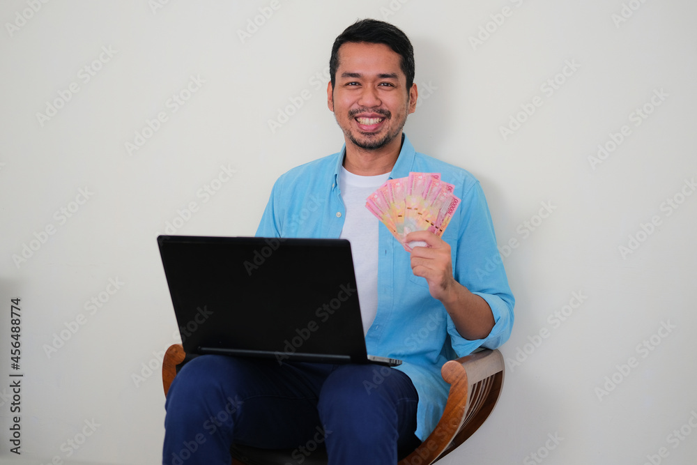 Adult Asian man sitting in a wooden chair while smiling happy showing his paper money Adult Asian man sitting in a wooden chair while smiling happy showing his paper money