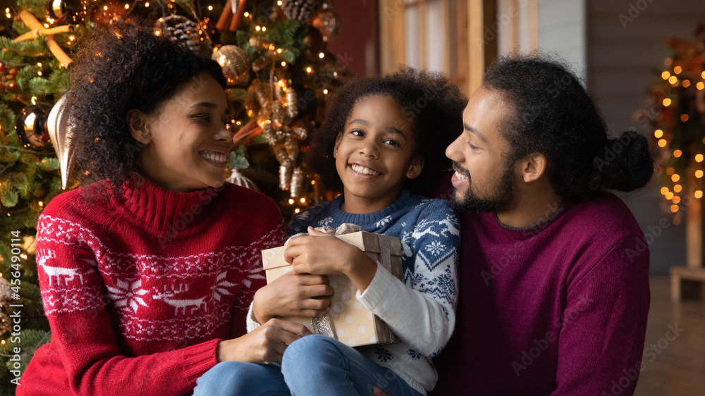 Fototapeta premium Smiling young African American family with teen daughter sit near fir tree celebrate New Year winter holidays together. Happy biracial parents with girl child open gifts on Christmas celebration.