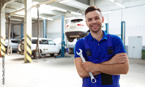 Confident handsome young and experienced car repair worker in work overalls posing against the background of lifted cars in a car service