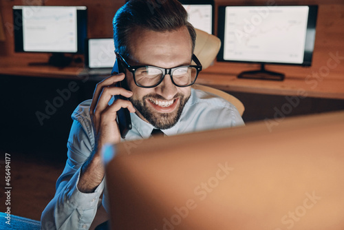 Happy young man talking on mobile phone and looking at computer while staying late in the office