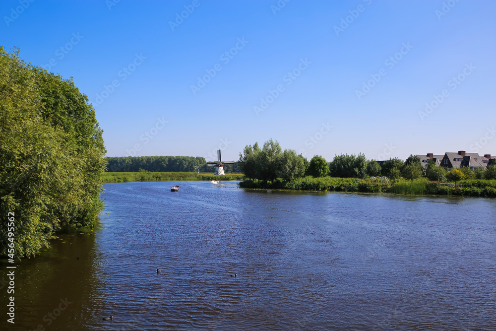 View on river Alblas with green trees, residential houses and windmill against blue summer sky - Alblasserdam, Netherlands