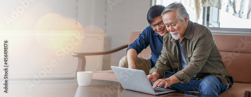 happy asian adult son and senior father sitting on sofa using laptop together at home . young man teaching old dad using internet online with computer on couch in living room . copy space banner