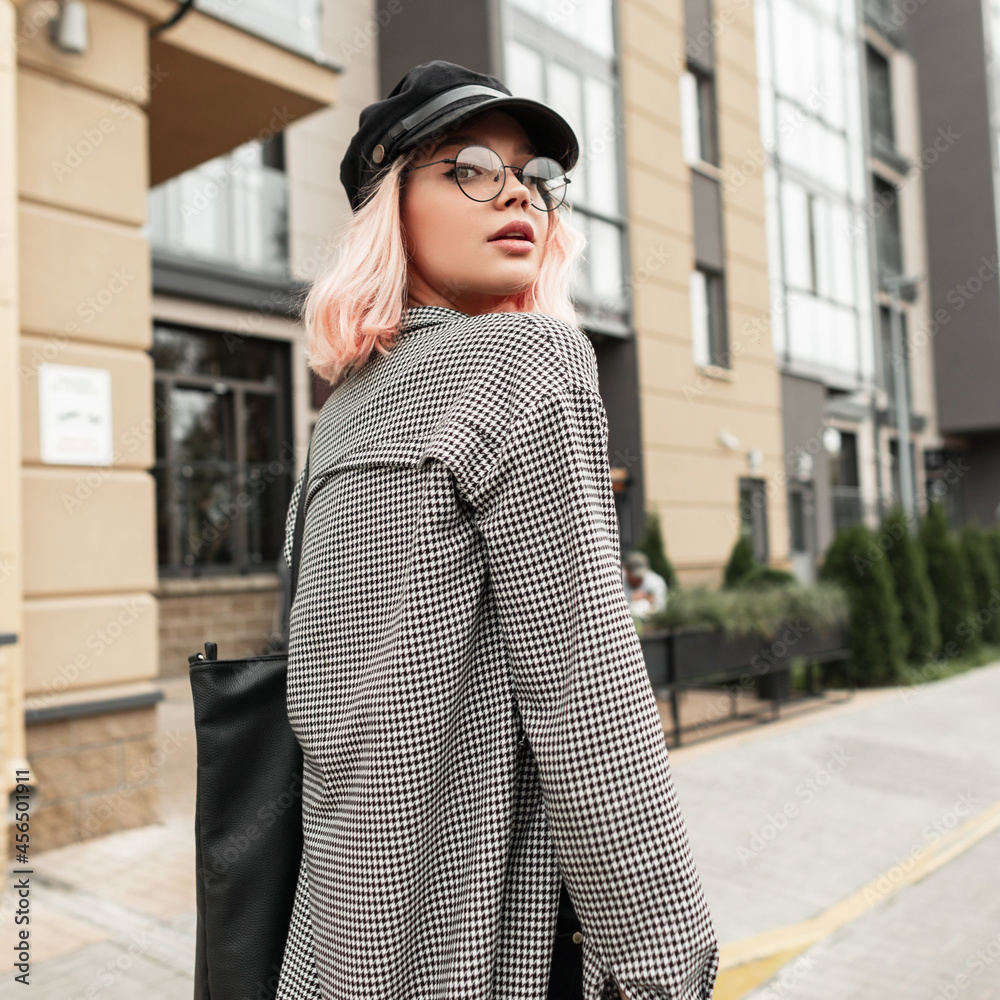 Fototapeta premium Cute young woman with stylish glasses and vintage hat with trendy casual shirt walking on the street near the building