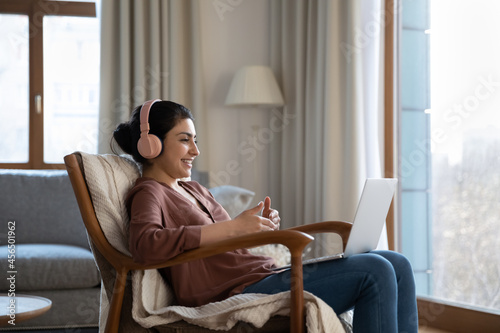 Cozy education. Smiling mixed race female in headphones sit in comfy chair hold laptop talk to teacher by video call repeat foreign language phrases. Young indian woman enjoy studying from home online