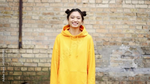 Slow motion of modern asian girl in streetstyle clothes, smiling and looking happy, laughing while standing outdoors on street against brick wall. Face of young candid woman