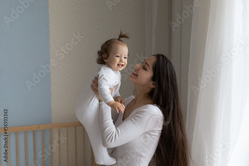 Canvas-taulu Happy new mom enjoying motherhood, maternity leave, holding, lifting cute funny toddler child at crib, laughing, talking to baby