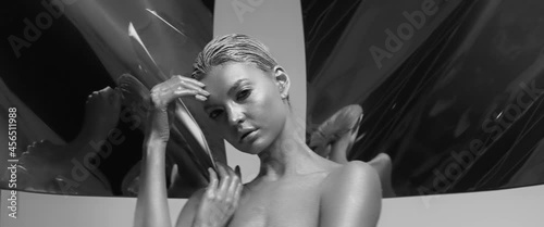 Young woman with skin and hair dyed silver posing emotionally and dancing in studio. Beautiful girl with trendy metallic makeup looks at camera, sensually moving. Anamorphic lens shot