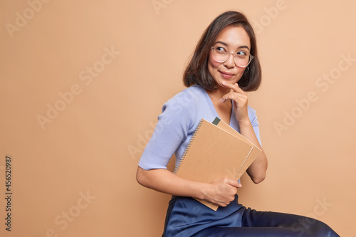 Thoughtful Asian female schoolgirl carries spiral notebooks returns back to school thinks how to improve her knowledge wears spectacles casual clothes sits indoor blank copy space on beige background