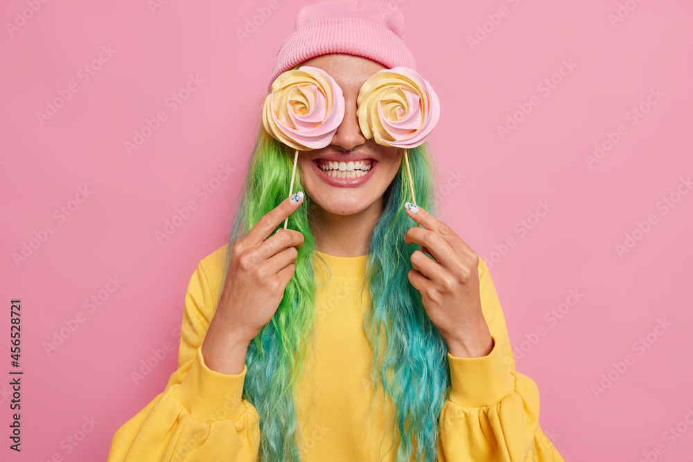 Photo of positive woman has colorful hair covers eyes with caramel candies smiles broadly has sweet tooth enjoys cheat meal day foolishes around wears casual clothes poses against rosy background Photo of positive woman has colorful hair covers eyes with caramel candies smiles broadly has sweet tooth enjoys cheat meal day foolishes around wears casual clothes poses against rosy background