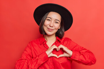 Gentle smiling Asian woman shows heart sign near chest I love you gesture wears black hat and shirt poses against vivid red background wishes happy valentines day. Romantic message compassion