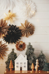 Stylish christmas little house, trees, golden lights on background of paper stars on white wooden wall. Festive decorated scandinavian room. Atmospheric hygge decor. Magic time. Merry Christmas!