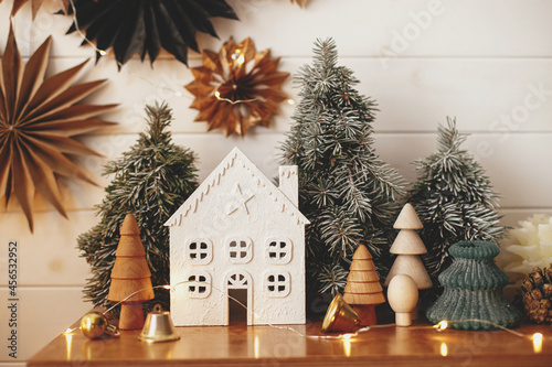 Stylish christmas little house, trees, golden lights on background of paper stars on white wooden wall. Atmospheric festive  decor in modern scandinavian room. Magic winter time. Merry Christmas!
