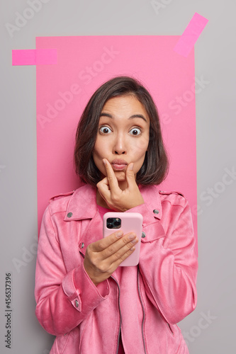 Surprised Asian woman keeps lips folded looks with stunned expression uses cellphone advertises new mobile application texts online dressed in jacket poses indoor. Technology and communication