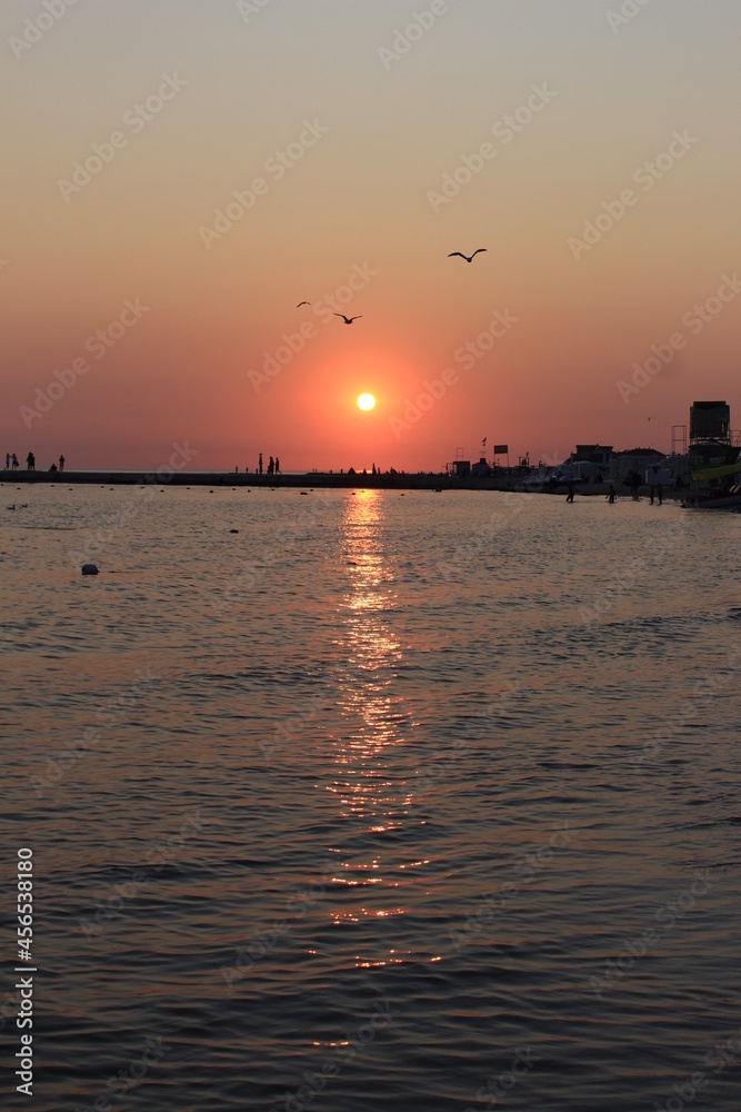 Fototapeta premium Sunset paints the sky and sea in shades of red. Seagulls fly against the backdrop of the setting sky and the setting sun. The sea pier is visible in the distance. The resting people watch the sunset