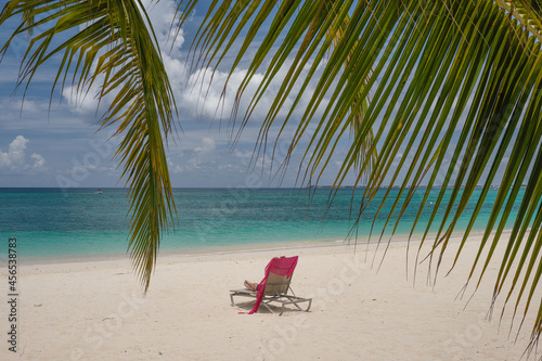 A sole chaise lounge chair sits on an empty beautiful clear and turquoise water beach on the Cayman Islands near a small grove of palm trees