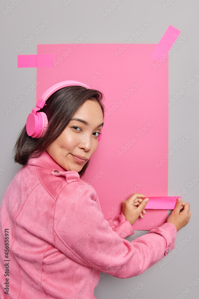 Back view of mysterious young Asian woman plasters pink sheet of paper on grey background to place your advertisement listens music wears jacket looks seriously at camera. Empty space for text Back view of mysterious young Asian woman plasters pink sheet of paper on grey background to place your advertisement listens music wears jacket looks seriously at camera. Empty space for text